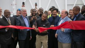 Photo of several men of different ethnicities, smiling, and holding a long red ribbon, one gentleman in the center is cutting the ribbon with scissors